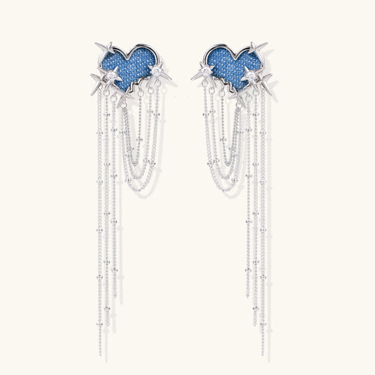 Long dangle earrings with blue denim heart shapes, silver star spikes, and multiple cascading chains on beige background.
