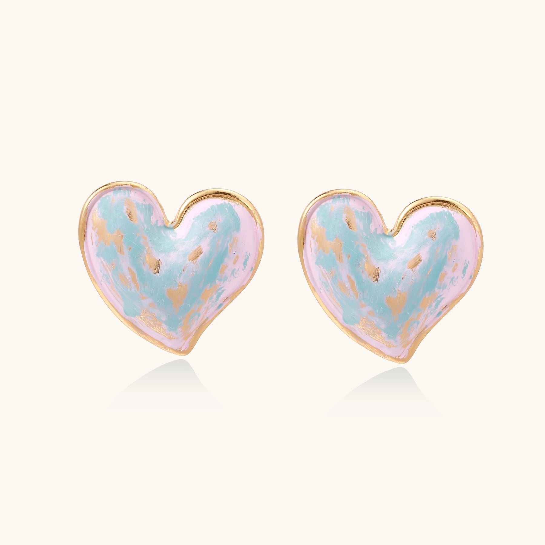 Heart-shaped stud earrings with iridescent blue-pink enamel finish and gold-plated brass frames against cream background